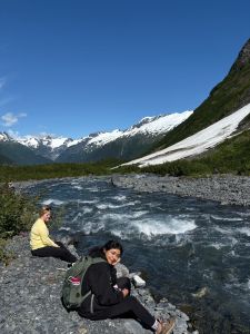 Two students sitting next to a rapidly moving river in Alaska with mountains in the background
